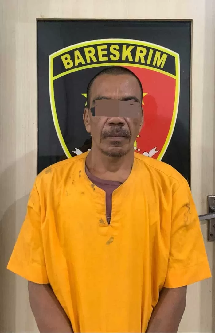 Seorang polisi berdiri di lokasi kejadian pembacokan tragis di Dusun Tanjung Mutiara, Inhil. Lokasi tersebut dipasangi garis polisi berwarna kuning. Beberapa warga sekitar tampak berkumpul dan menyaksikan tempat kejadian dengan wajah cemas dan sedih